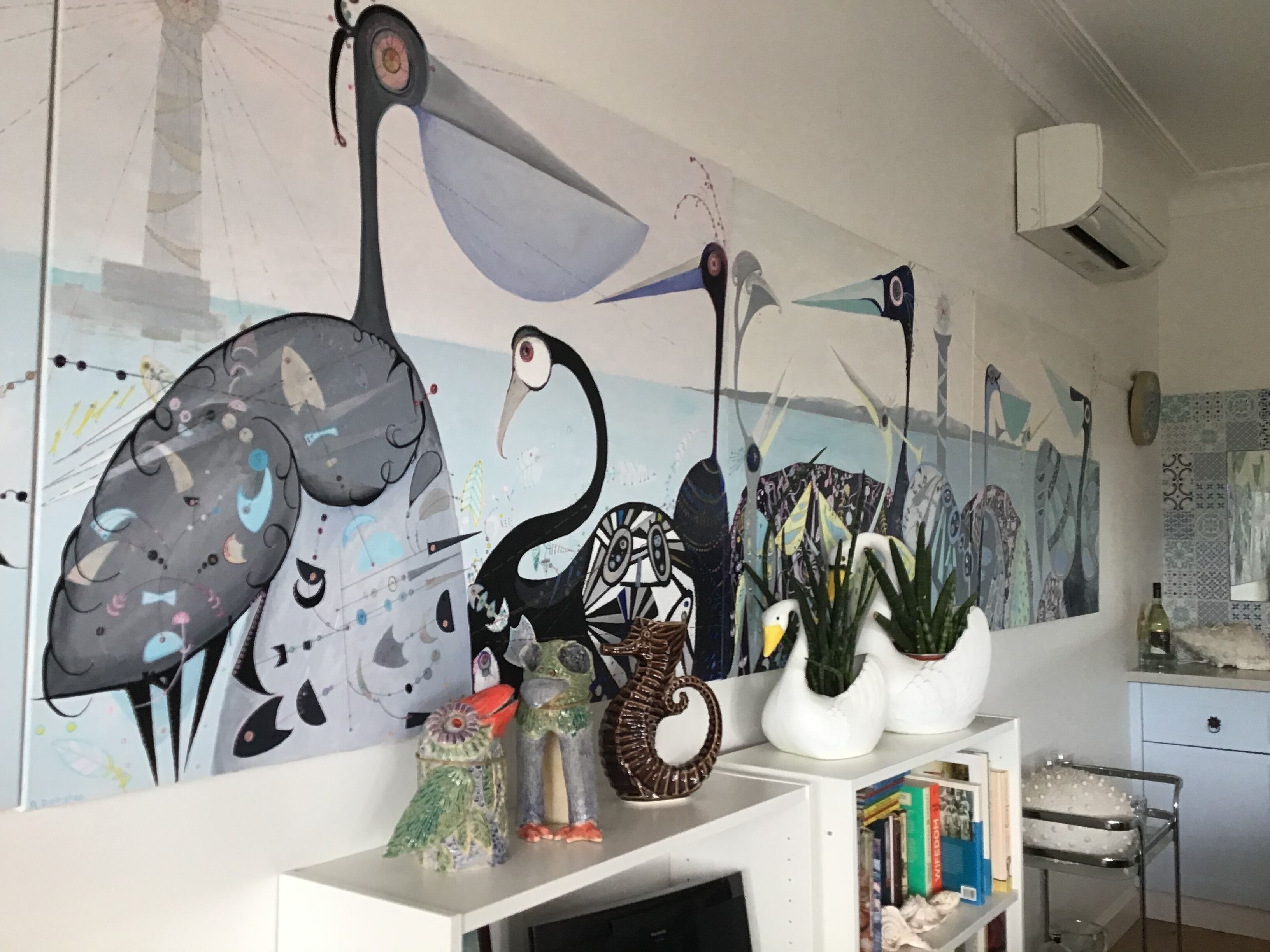 Een kamer met een grote abstracte muurschildering, een wit schap met boeken en decoratieve objecten, en een kleine tafel met een vaas en een fles.