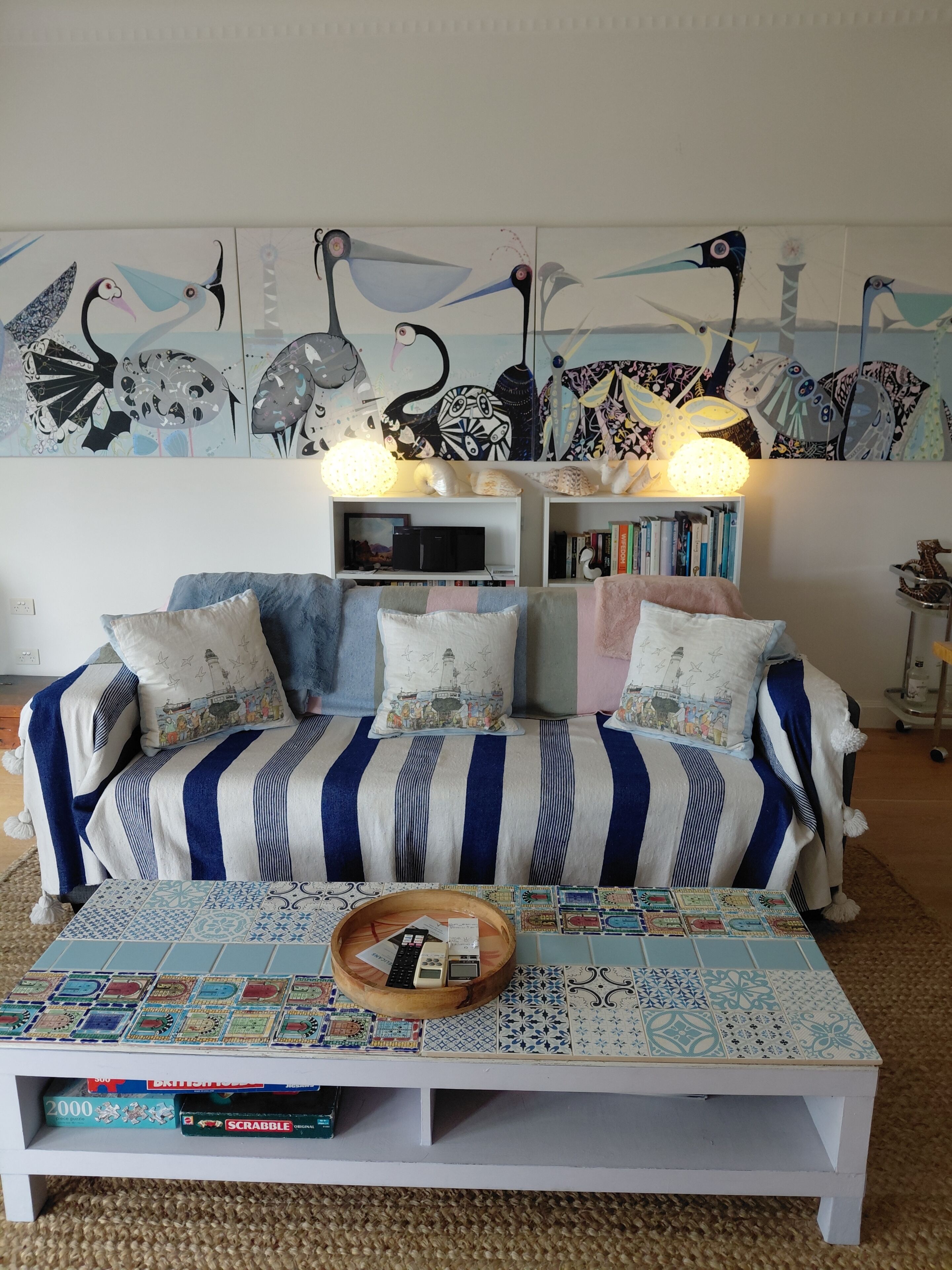 A living room with a striped sofa, a coffee table with board games, a bookshelf, and a wall mural.