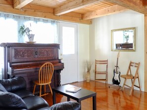 A living room with a wooden piano, a black leather sofa, two wooden chairs, a guitar, and a table with a magazine.