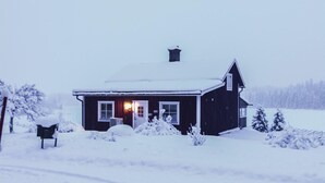 Una casa cubierta de nieve con una puerta principal y ventanas visibles, rodeada de árboles nevados y una cerca.