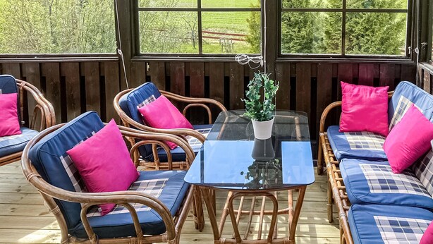 Un espacio al aire libre techado con mobiliario de mimbre y cojines, una mesa con cubierta de vidrio y vistas a un paisaje verde a través de amplios ventanales.