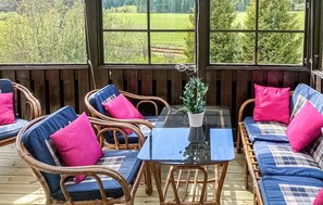 Un espace extérieur aménagé, abrité et doté de mobilier en osier et de coussins, d’une table à plateau en verre, et offrant une vue sur un paysage verdoyant grâce à de grandes fenêtres.