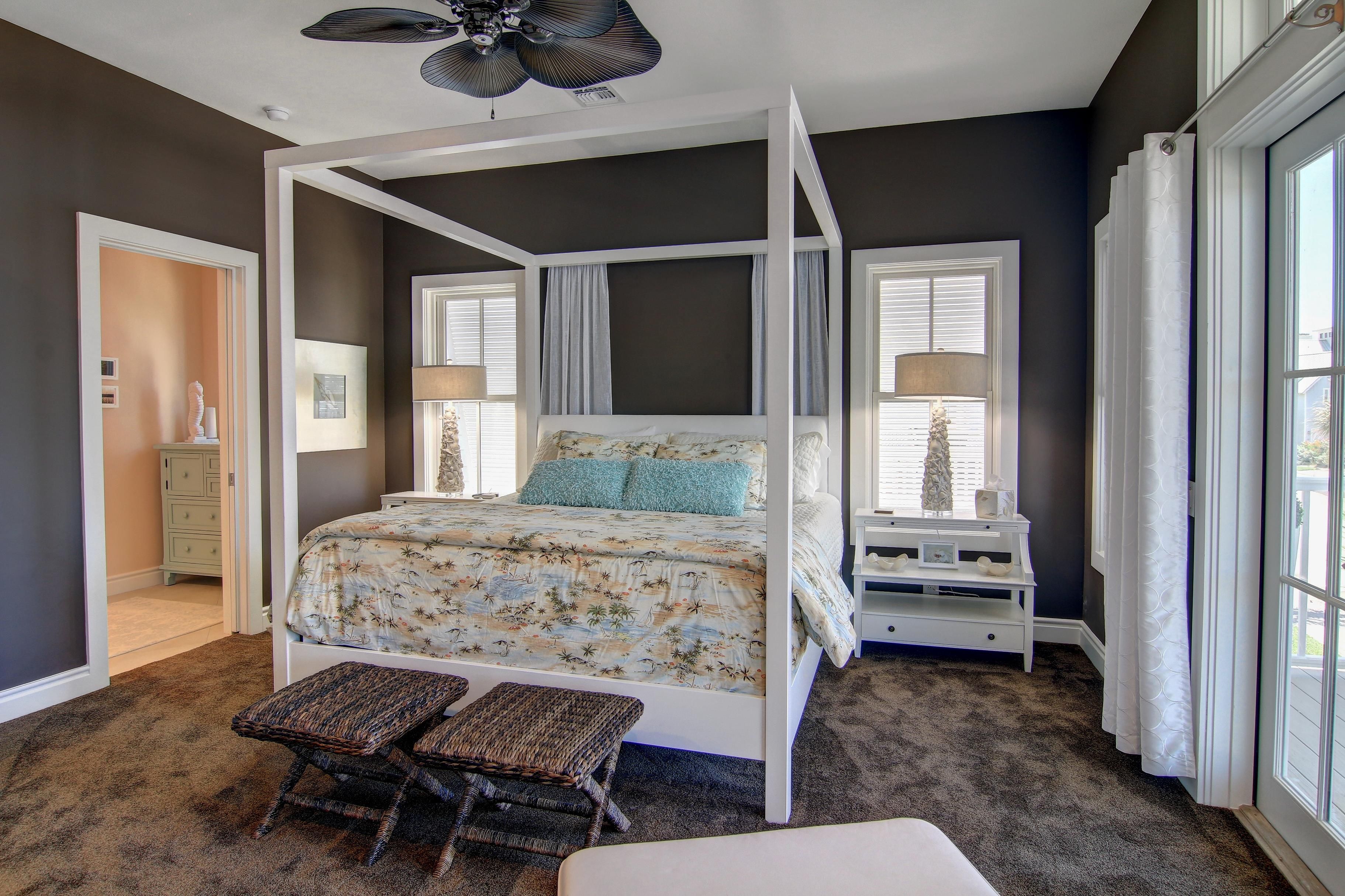 A four-poster canopy bed with a floral bedspread, two wicker chairs, a white nightstand with a lamp, and a dresser with a mirror.
