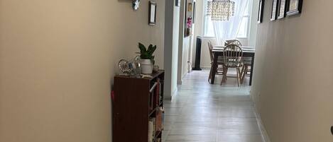 A hallway with a marble floor, framed pictures on the walls, a bookshelf, and a dining area in the distance.