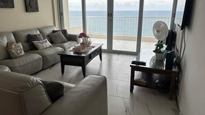 A living room with a sofa, coffee table, and a view of the ocean through large windows. - Luquillo, 13th Floor with Ocean Front! (Luquillo)