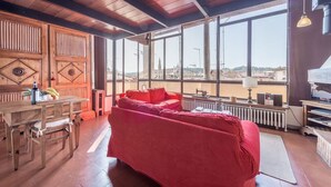 Ein Zimmer mit Holzboden, einem roten Sofa, einem Holztisch mit Stuhl, einem Heizkörper und großen Fenstern mit Blick auf eine Stadtlandschaft.