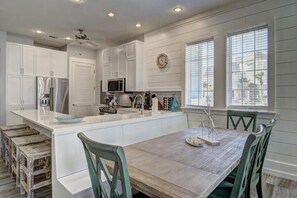 A modern kitchen with white cabinetry, a central island, and a dining area with a wooden table and chairs. - Pet-Friendly Condo w/ Beach Fun Decor & Resort Amenities@Cinnamon Shore (Port Aransas)