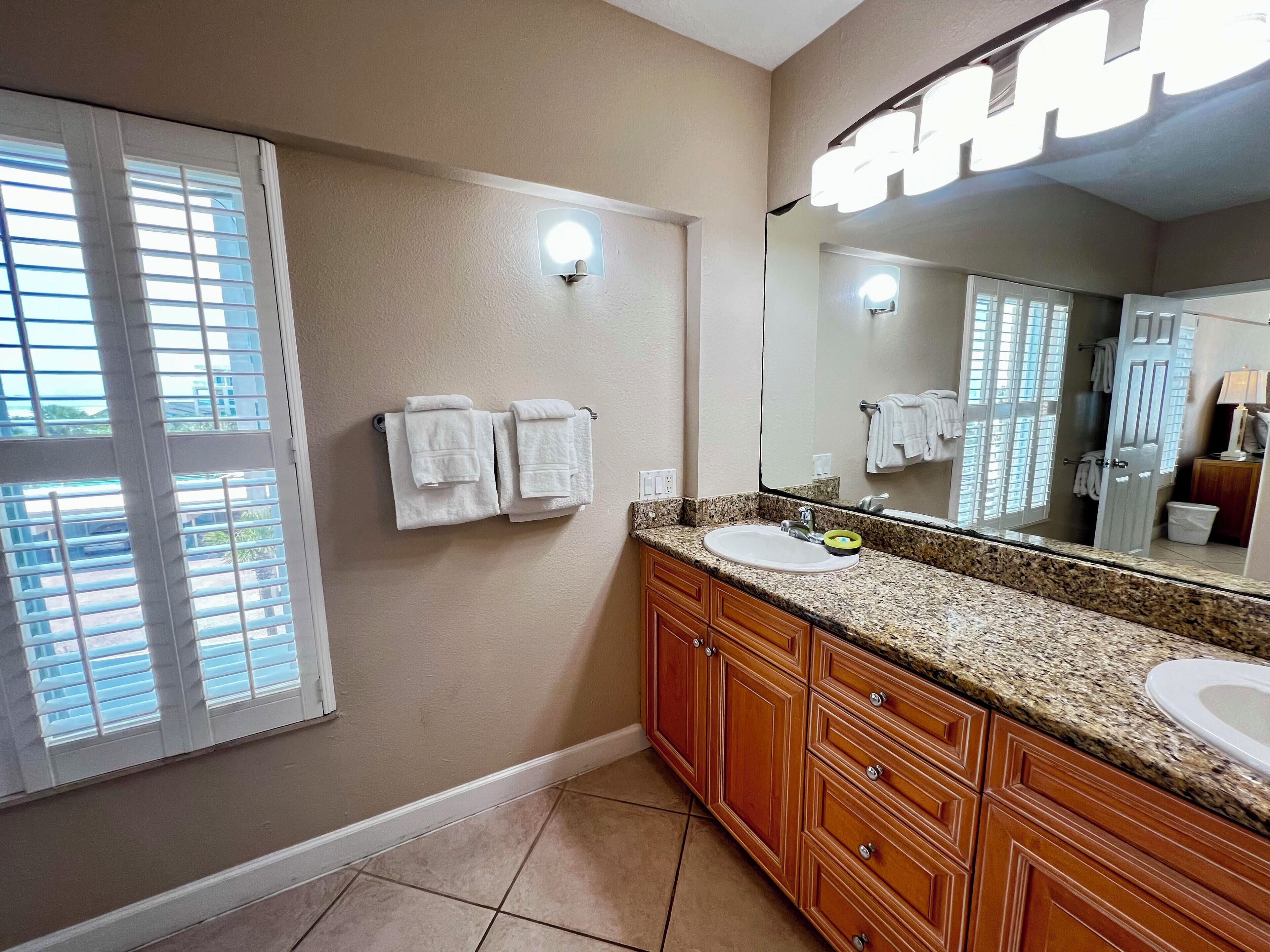 A bathroom with a granite countertop, wooden cabinets, a large mirror, and a window with blinds.