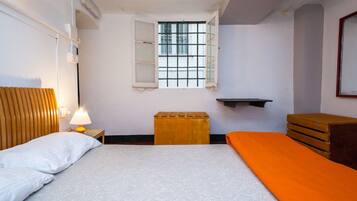 Una habitación pequeña y limpia con una cama, una mesita de noche, una ventana y un cofre de madera.