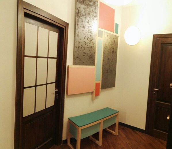A hallway with a wooden door, a bench, and a wall with a light fixture and a geometric art piece. - Ostellin Genova Hostel (Genoa)