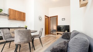 A modern living room with a grey sofa, two armchairs, a coffee table, and a TV. There is a wooden cabinet above the kitchen counter, and a small plant on the counter. - 1 bedroom nice apartment in Podgradina (Podgradina)