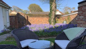 Een terras met rieten meubilair en een tafel, omgeven door een tuin met paarse bloemen en een bakstenen muur.