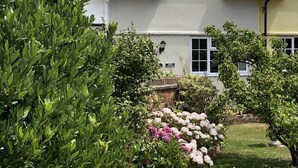 Een charmant landhuis met een bakstenen dak, witte muren en een verzorgde tuin met diverse struiken en bloeiende planten.
