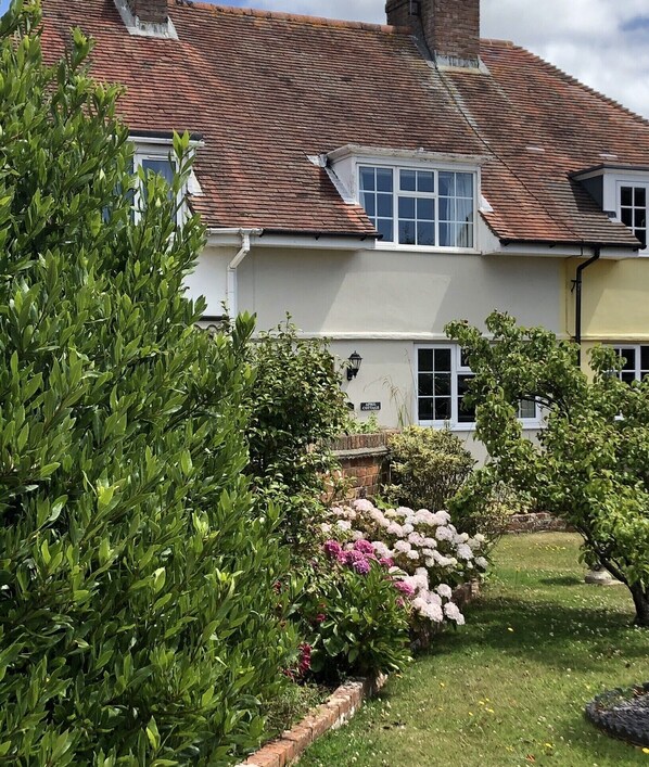 Une maison de campagne traditionnelle avec un toit en briques, des murs blancs et un jardin bien entretenu, agrémenté de divers arbustes et de plantes à fleurs.