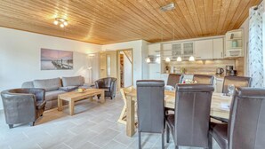 A modern living room with a wooden ceiling, a sofa, a coffee table, and a dining area with chairs and a table. - Ferienhaus 4 Altefähr (Altefähr/Rügen)
