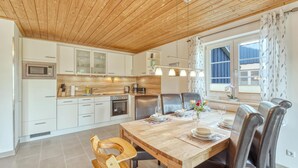 A modern kitchen with white cabinets, a wooden ceiling, and a dining area with a wooden table and leather chairs. - Ferienhaus 4 Altefähr (Altefähr/Rügen)