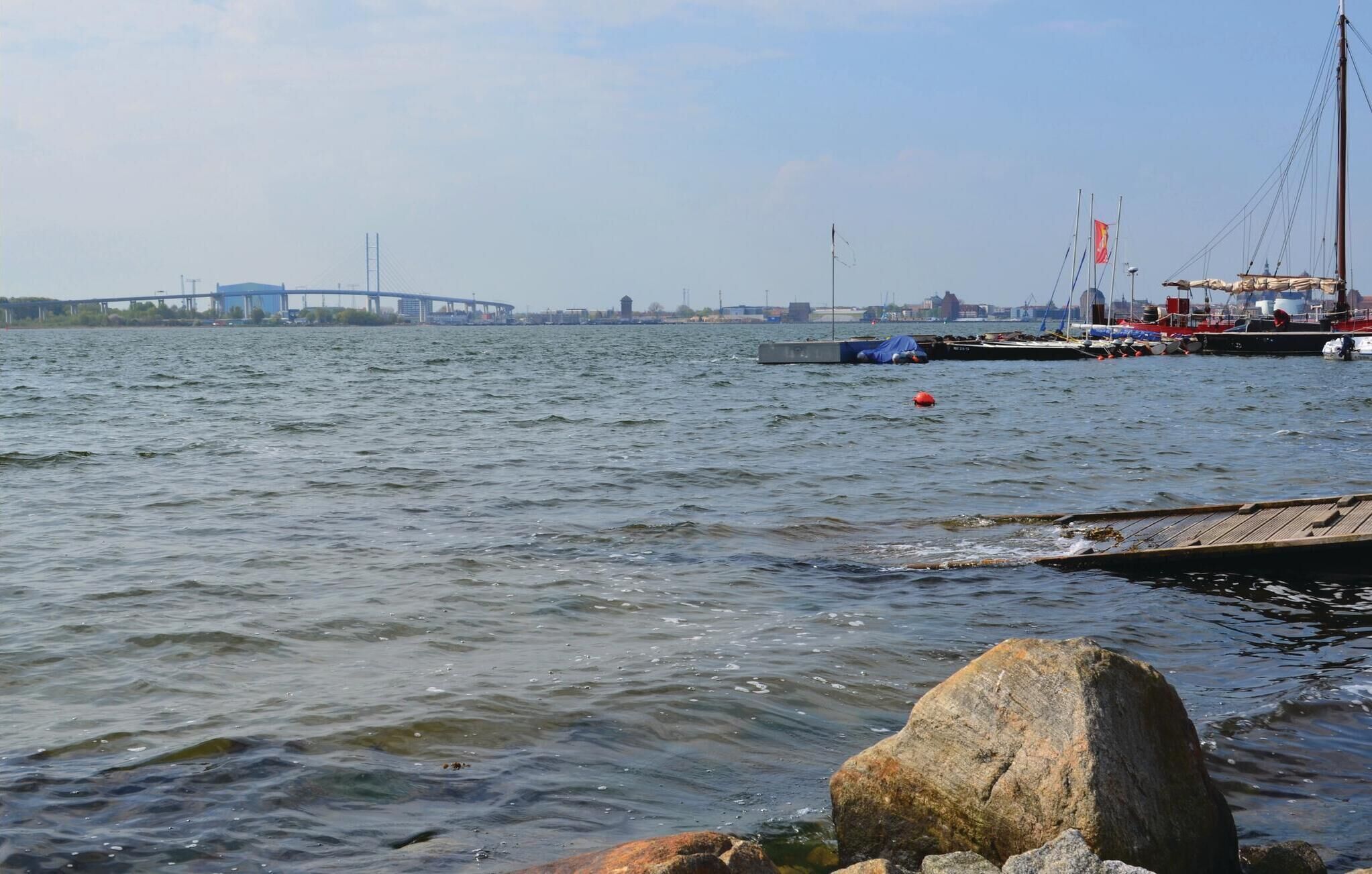 Eine Uferlandschaft mit einer Brücke im Hintergrund, ruhigem Wasser und einem Steg mit Booten.