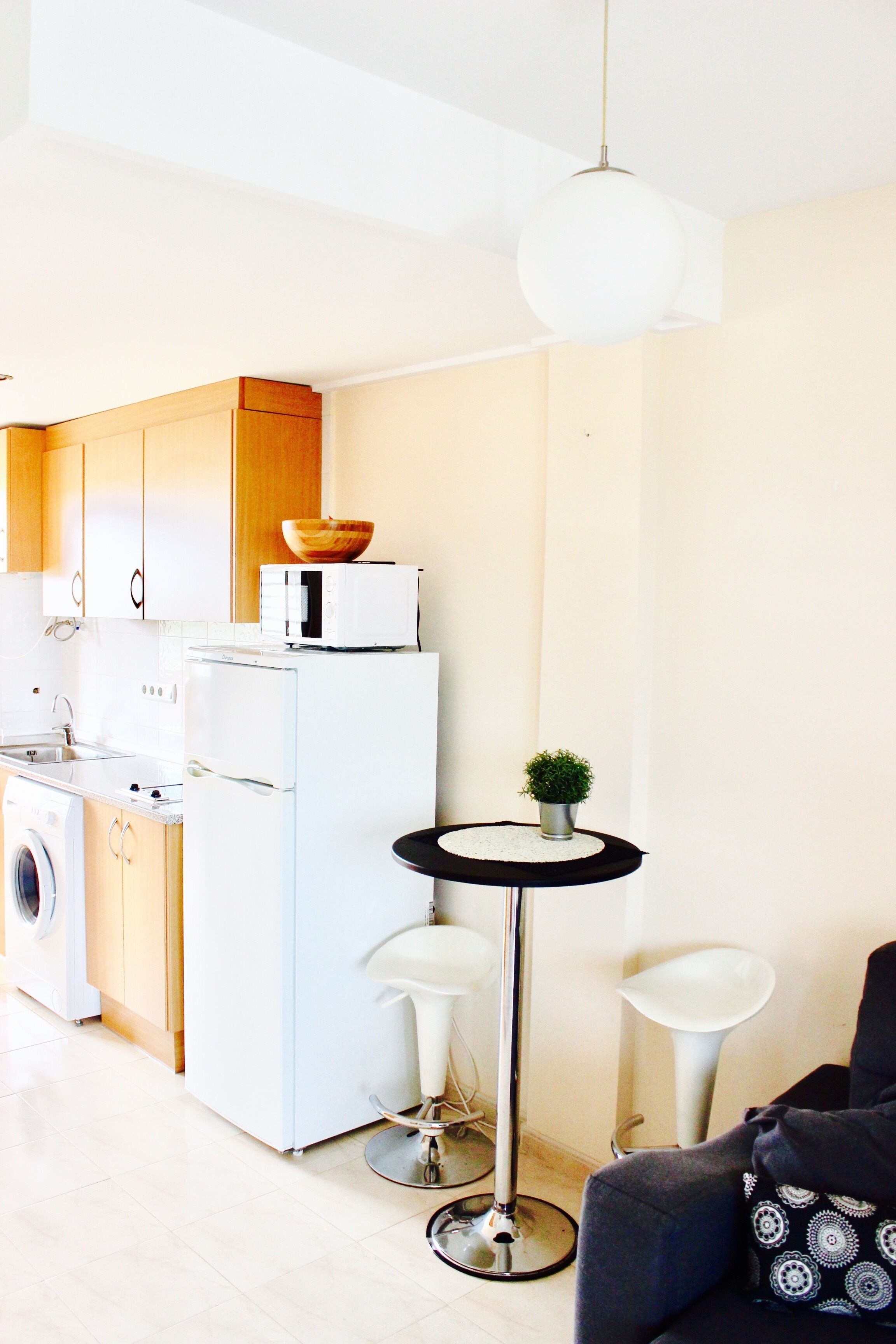 A compact kitchen with a washing machine, a refrigerator, and a microwave. There is a small round table with a potted plant and two stools. A modern pendant light hangs from the ceiling.