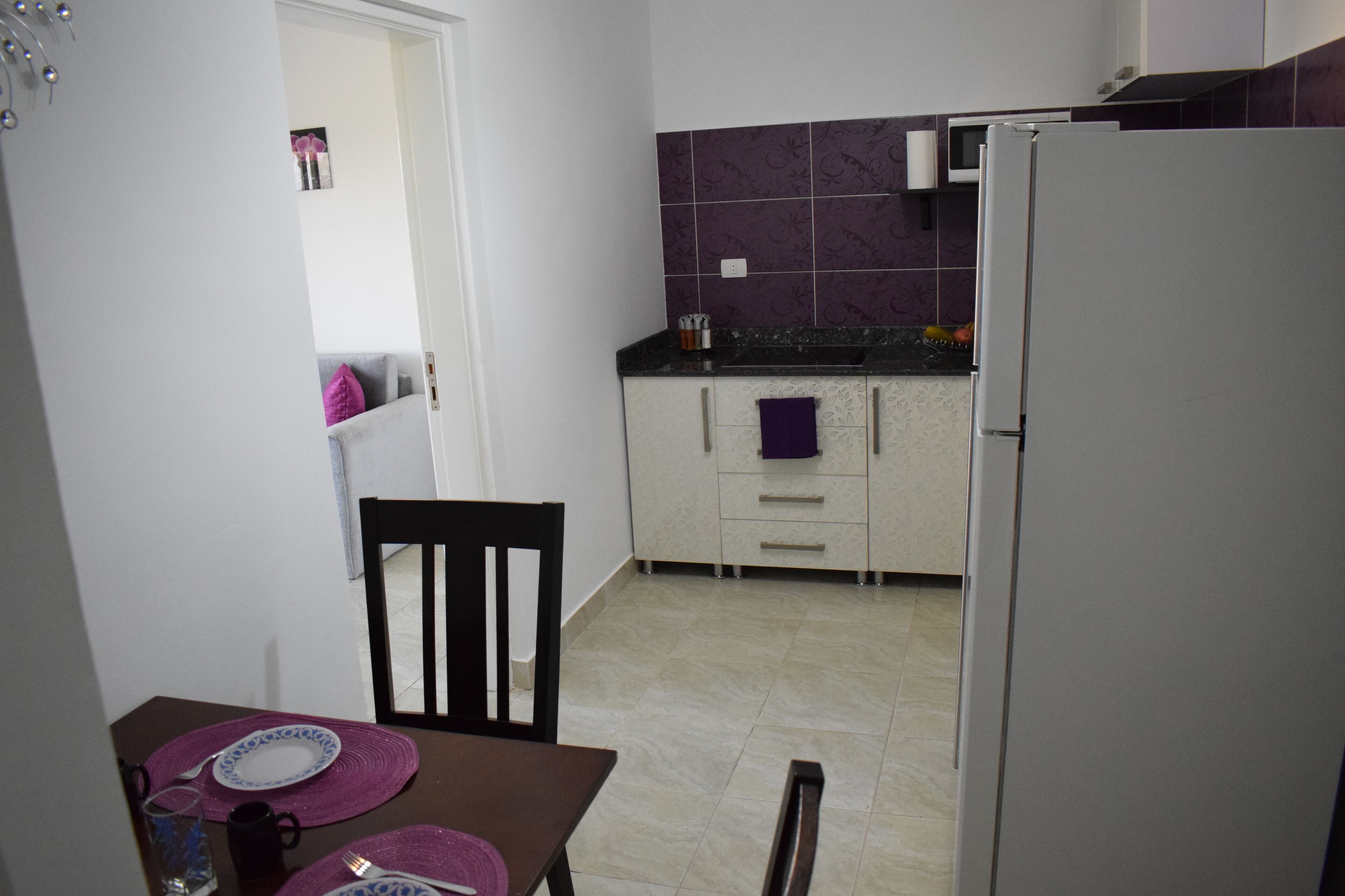 Cocina compacta con refrigerador, microondas y lavaplatos. Hay una mesa de comedor con una silla y un mantel individual morado.