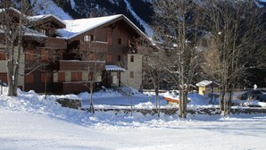 Un edificio de madera de varios pisos con balcones, rodeado de nieve en el suelo y árboles, con montañas al fondo.