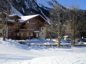 A multi-story wooden building with balconies, surrounded by snow-covered ground and trees, with mountains in the background.