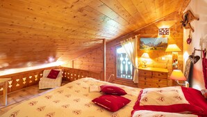 Un dormitorio acogedor en el ático, con techo de madera, cama con ropa de cama roja y blanca, mesita de noche con lámparas y ventana con cortinas.