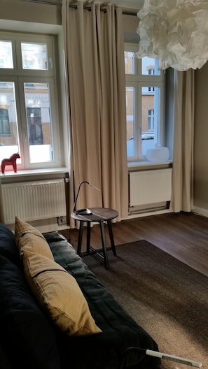 Una habitación con un sofá negro, una mesita y una silla. Hay ventanas con cortinas, un radiador y vistas a edificios.