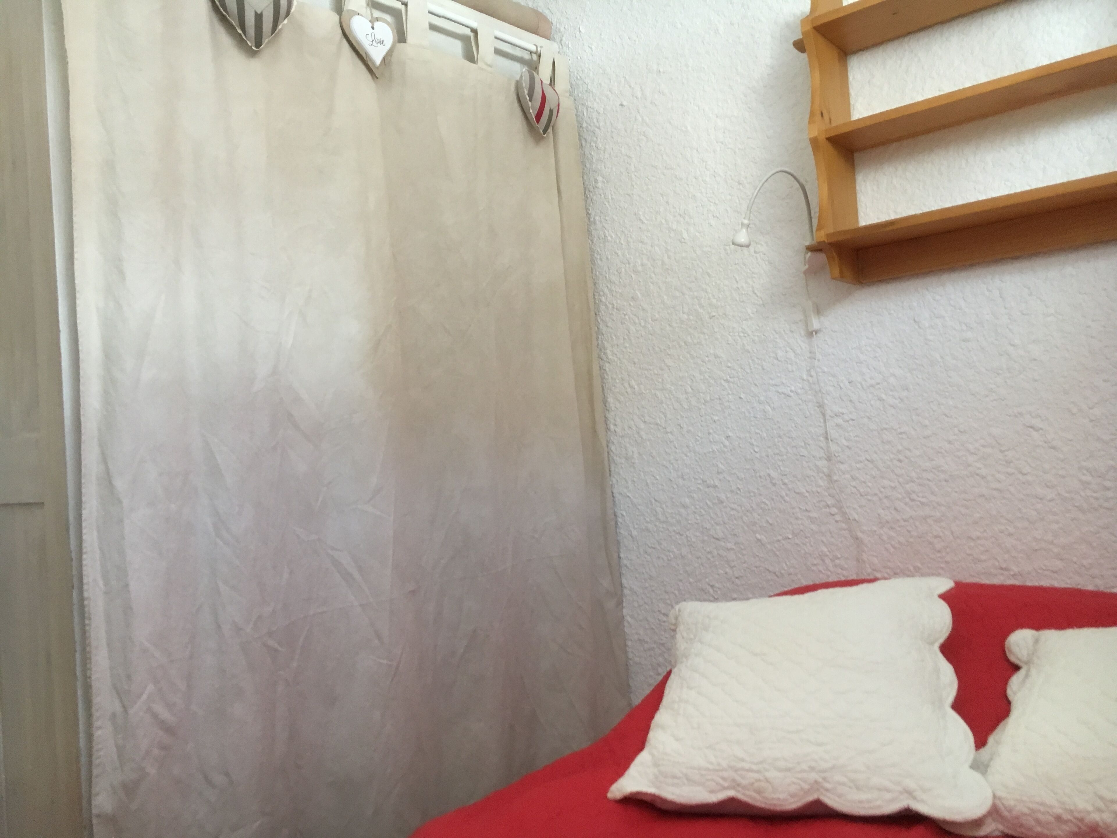 Une chambre à coucher avec un lit, une couverture rouge, un oreiller blanc, une étagère en bois et un mur blanc à texture.