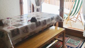 A cozy room with a wooden bench, a table with a patterned tablecloth, a view of a wooden deck and trees outside. - T2 / 3, heart of resort, bottom of slopes, parking, 30m3 + 100m2 terrace (Mont-de-Lans)