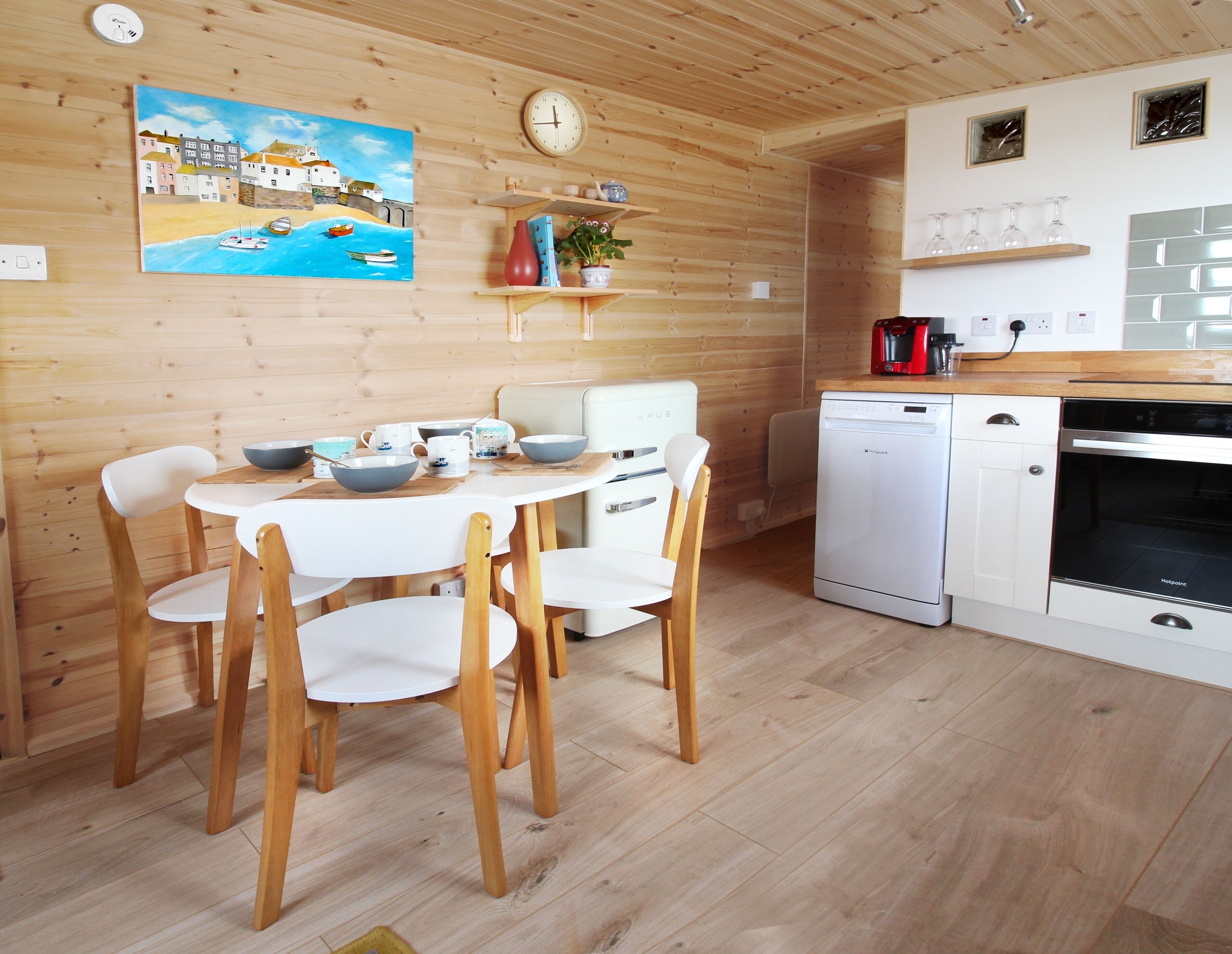 A cozy kitchen with wooden walls, a dining table with chairs, a coffee machine, and a dishwasher.