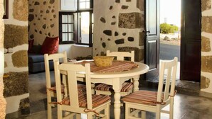 A rustic stone-walled dining area with a round table and six chairs, a wooden ceiling beam, and a view through an open door. - Casa de Los Olivos (Macher)