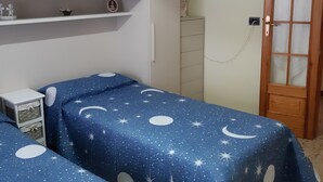 Et soveværelse med to senge med blåt sengetøj med et stjernenat-mønster, en hvid garderobe, en trædør og et lille natbord med en lampe.