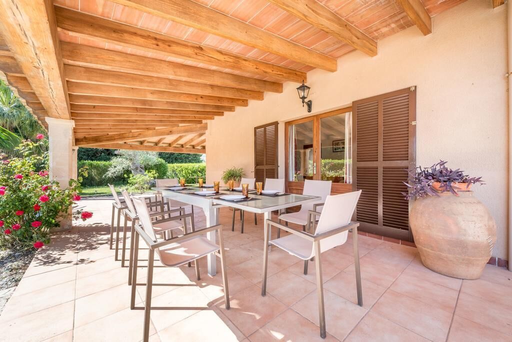 Un espace extérieur abrité, aménagé pour les repas, avec une table et des chaises, un grand bac à fleurs et une vue sur le jardin.