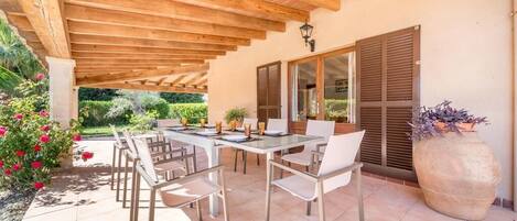 Un espace extérieur abrité, aménagé pour les repas, avec une table et des chaises, un grand bac à fleurs et une vue sur le jardin.