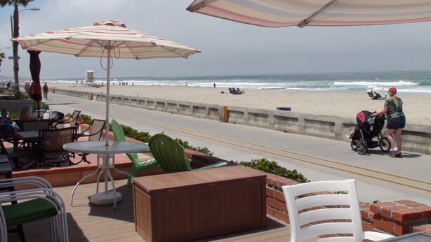 A beachside patio with outdoor seating, a view of the ocean, and a lifeguard tower in the background.