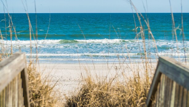 Blick auf einen Strand mit Wellen, einer Holzpromenade und hohem Gras im Vordergrund.