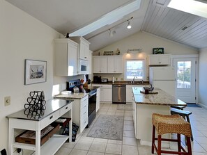 Une cuisine spacieuse avec des meubles blancs, un grand îlot central et un coin repas agrémenté d’une chaise en osier.