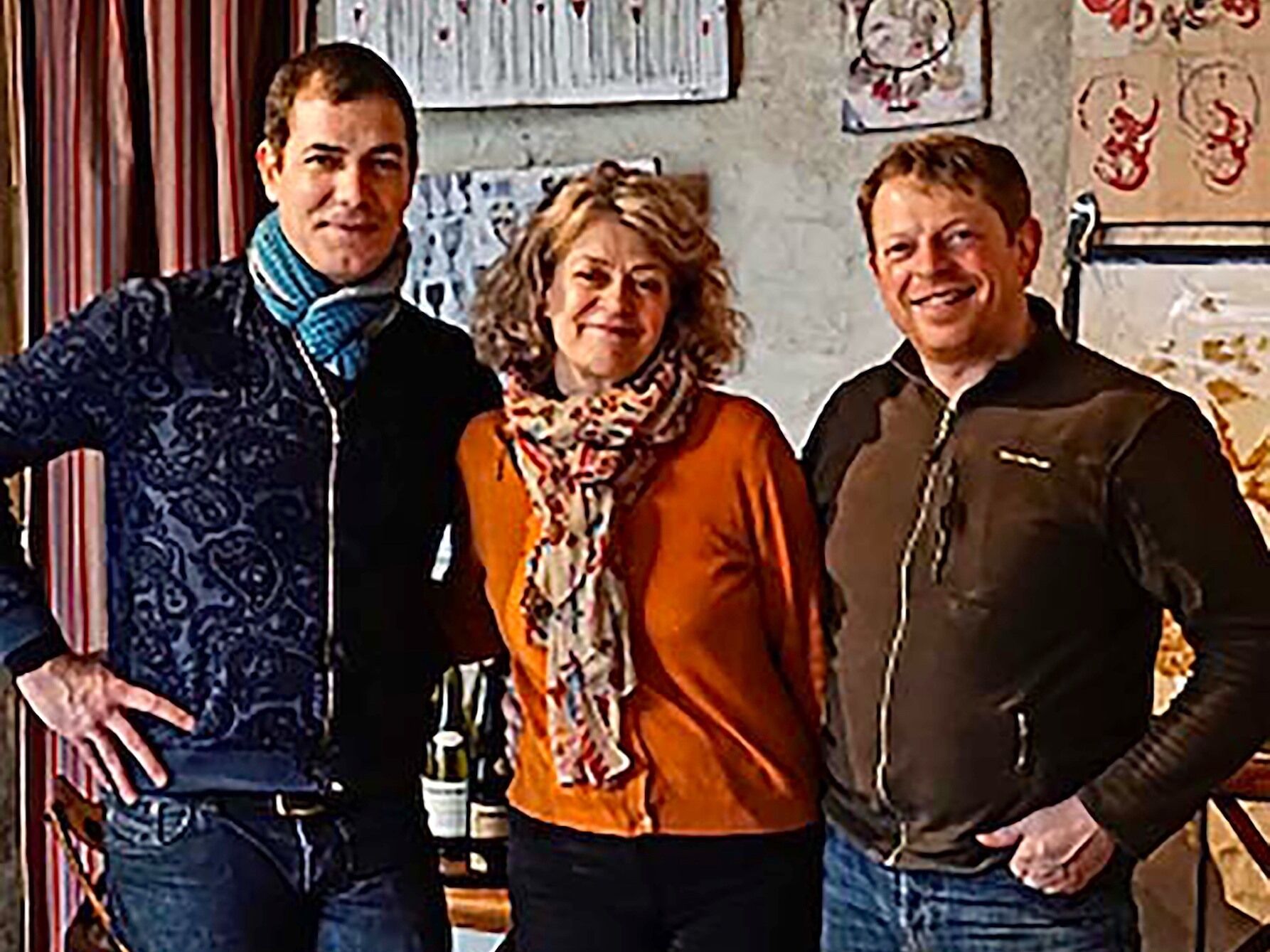 Three people standing indoors with a wall featuring various framed items and a shelf with bottles.