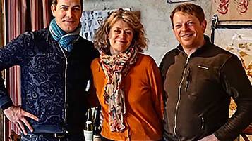 Three people standing indoors with a wall featuring various framed items and a shelf with bottles.