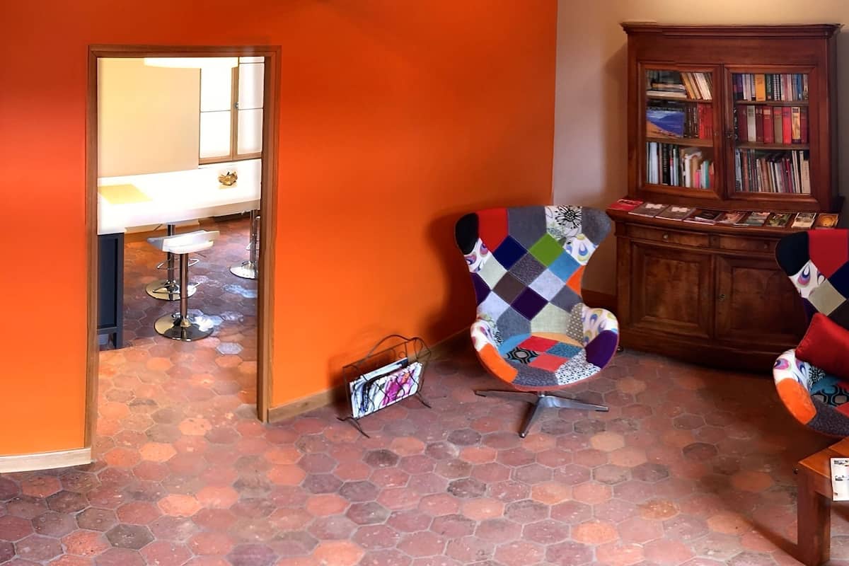 Ein Zimmer mit orangefarbenen Wänden, einem sechseckig gefliesten Boden, einem farbenfrohen Sessel, einer Holzhocker mit Büchern und einem weißen Tisch mit einer Lampe.