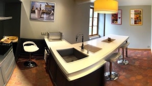 A modern kitchen with a central island, a built-in sink, and a microwave. There is a white chair and a bar stool near the island. - Les Temps Perdus (Prehy)
