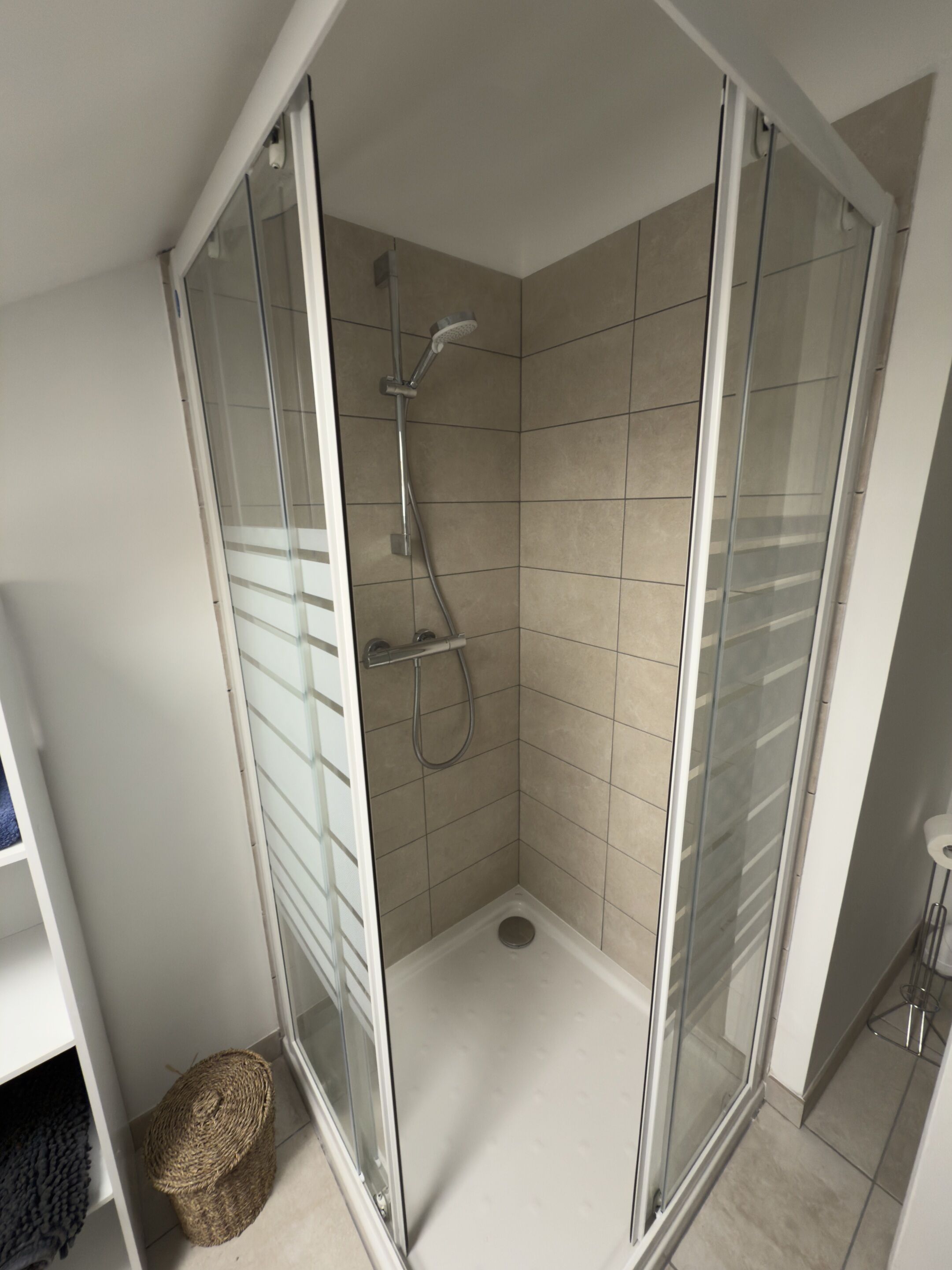 Une douche fermée par des parois en verre, dotée d’un pommeau de douche à main et d’un drain au sol.