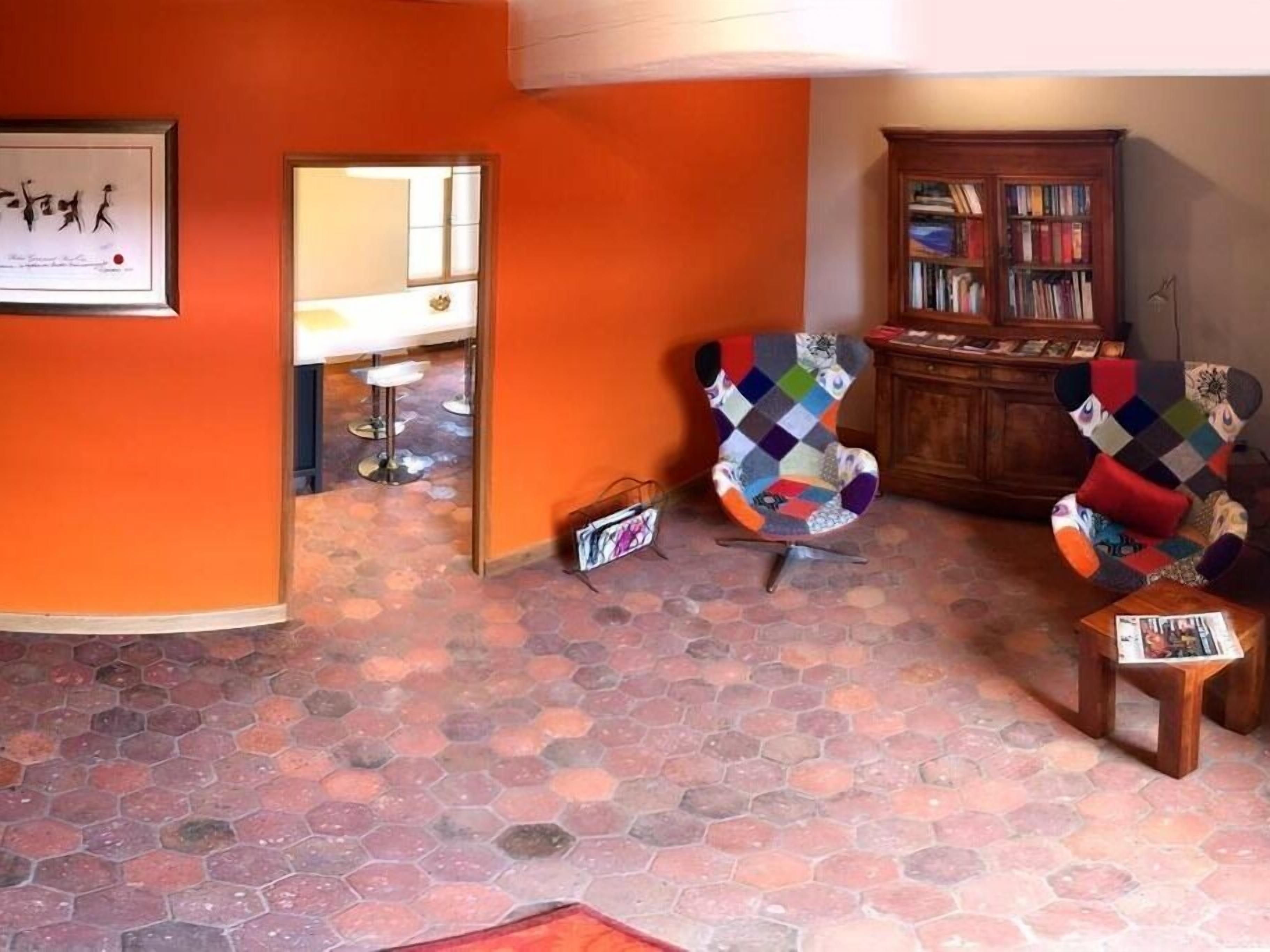 Ein Zimmer mit orangefarbenen Wänden, einem sechseckig gefliesten Boden, einem farbenfrohen Sessel, einer Holzkommode mit Büchern und einem gerahmten Bild an der Wand.