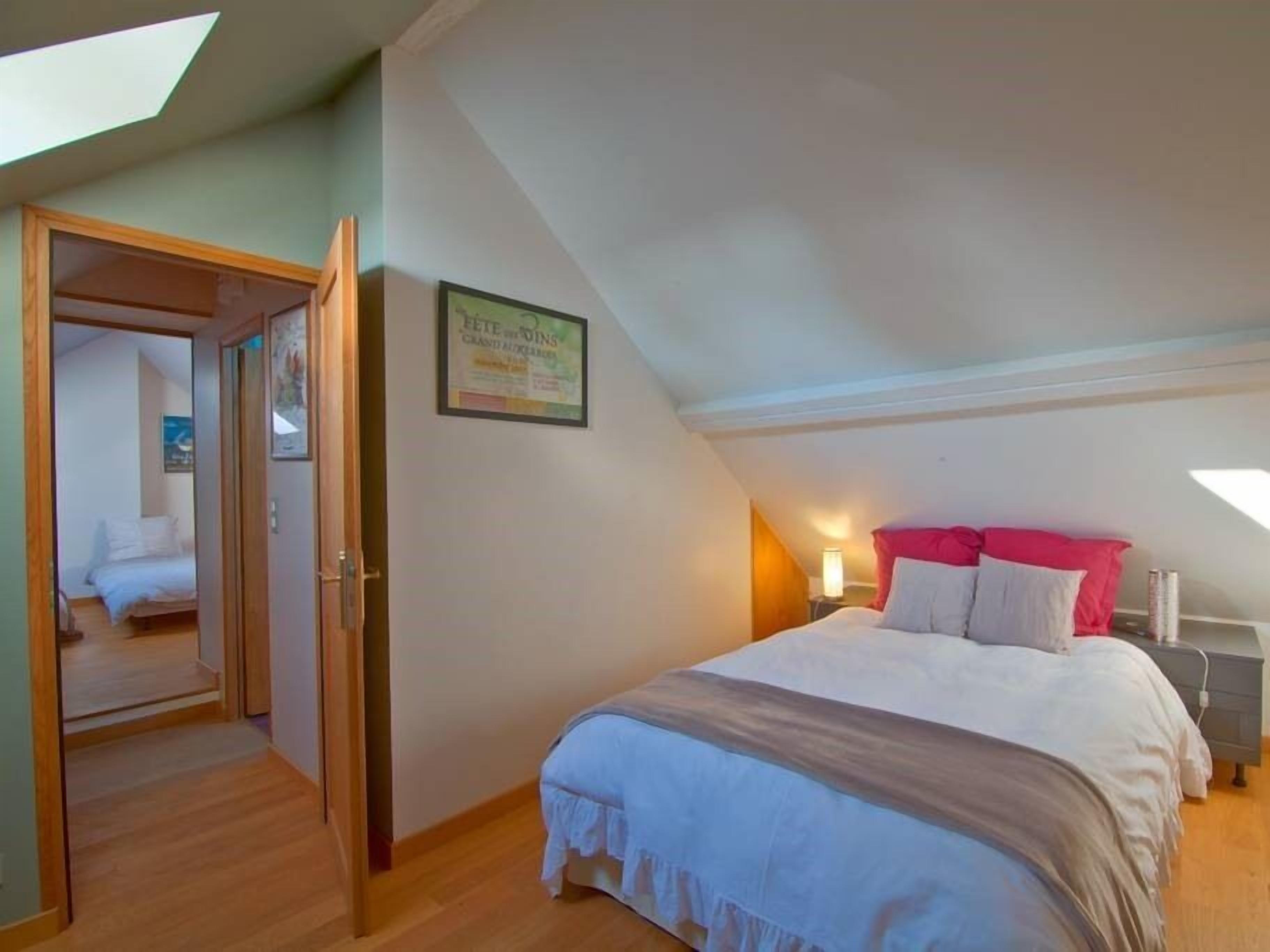 Een slaapkamer met schuine wand, een bed met wit en rode beddengoed, een nachtkastje en een wandlamp.