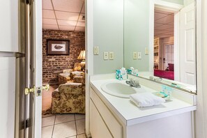 A bathroom with a white sink, a large mirror, and a door leading to a room with a brick wall and a lamp. - Intimate Getaway for Two, Steps from Forsyth Park by Lucky Savannah (Savannah)