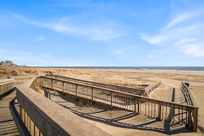 Ein hölzerner Steg führt zu einem Sandstrand unter blauem Himmel.