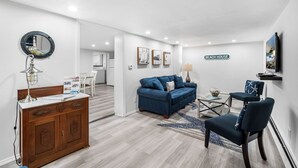 A modern living room with a blue sofa, a wooden cabinet, a round mirror, and a 'BEACH HOUSE' sign.