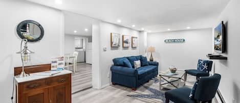 Una sala moderna con un sofá azul, un aparador de madera, un espejo redondo y un cartel que dice 'BEACH HOUSE'.