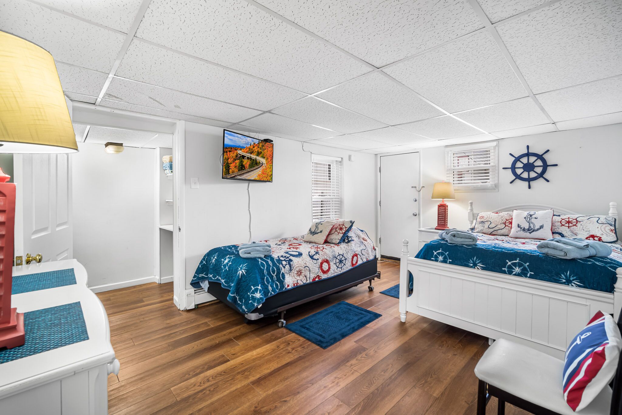 A bedroom with two beds, nautical-themed bedding, a mounted TV, and a ship's wheel decoration.