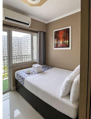 Ein Hotelzimmer mit einem Bett, einem Gemälde, einer Klimaanlage und Blick auf Gebäude durch ein Fenster.